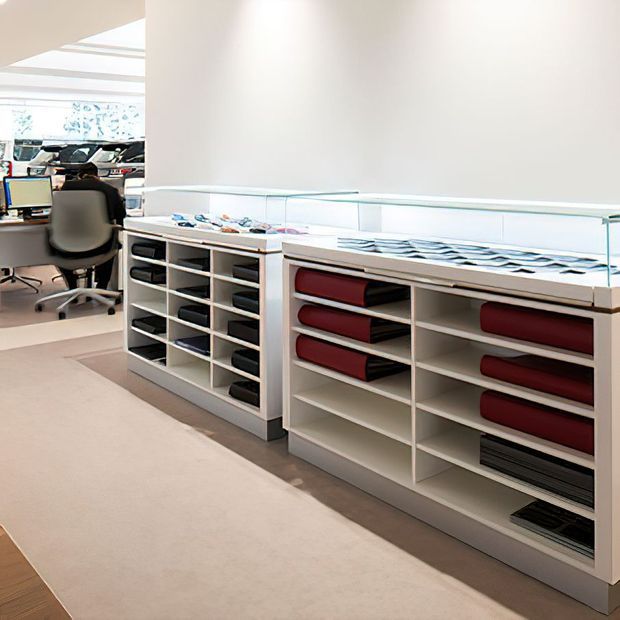 Bright, contemporary office or commercial workspace with long white built-in open shelving and display storage units. The shelving contains neatly arranged folders, binders, and documents, with glass-topped display counters above. A workstation with computer monitors and an office chair is visible in the adjoining area. The space has pale carpeting, white walls, abundant daylight, and a clean, highly organized fit-out.