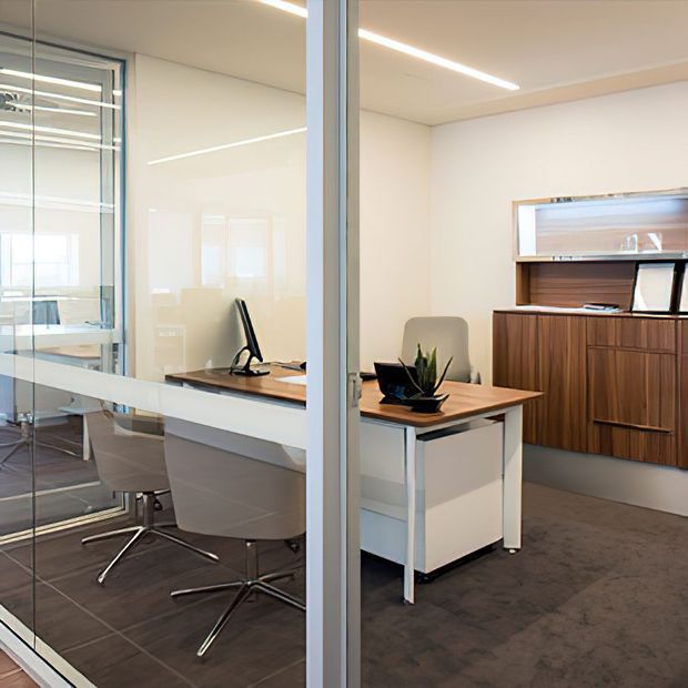 Modern office interior viewed through a glazed partition, featuring a compact workstation with a wood-topped mobile desk, upholstered task chairs, integrated cabinetry, and a dark carpeted floor. Linear recessed ceiling lighting and extensive internal glazing create a bright, professional workspace.