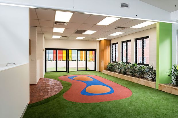 Bright commercial or institutional interior featuring an open indoor recreation or play area with artificial green turf, colourful red and blue patterned flooring, a curved timber-look section, full-height glazed doors, windows with blinds, planter beds, and a suspended acoustic ceiling with fluorescent lighting. A vivid green feature wall adds colour to the space.