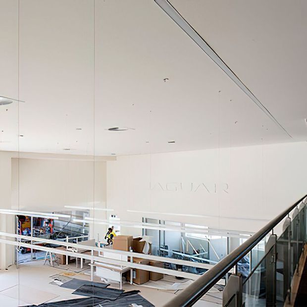 Bright, spacious contemporary commercial interior viewed from an upper-level stair or mezzanine. The image shows a large white suspended ceiling with recessed lighting, glass balustrades, white structural surfaces, benches, and work or retail areas below. A diagonal stair handrail dominates the foreground, while partial wall lettering and interior activity suggest a showroom, gallery, or office-like commercial space.