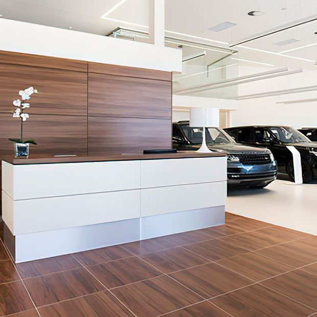 A bright, contemporary automotive showroom or dealership interior featuring a large white reception counter with integrated drawers, a horizontal dark timber wall panel behind it, and multiple black and dark-coloured vehicles displayed in the background. The space has a white ceiling with recessed lighting, exposed services, reflective surfaces, and a dark timber-look tiled floor.