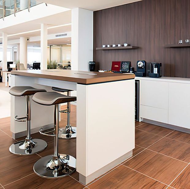 Bright, open-plan contemporary kitchen with a compact island breakfast bar, two sculptural adjustable stools, white cabinetry, a dark timber-look feature wall, integrated coffee appliances, and warm timber-toned tiled flooring. A glazed balustrade and open living area are visible in the background.