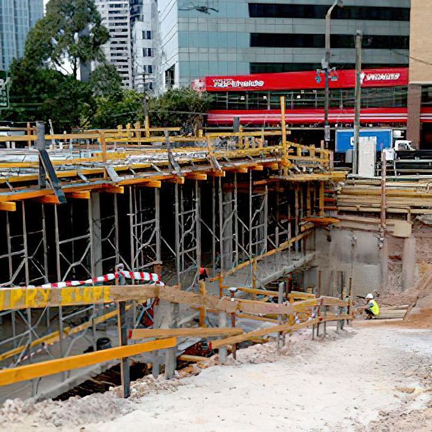 Urban construction site showing a deep excavated foundation or basement under construction, with extensive timber formwork, steel scaffolding, temporary barriers and structural supports. Multi-storey commercial and residential buildings are visible in the background, including a prominent red retail storefront.