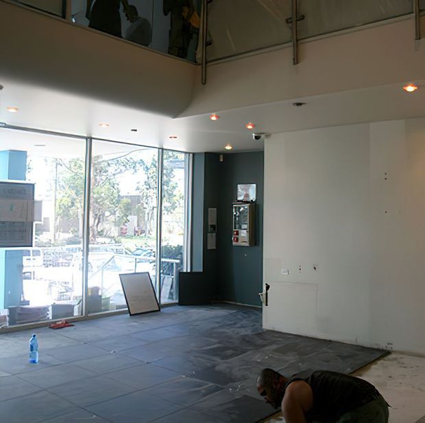 A partially finished, double-height commercial or retail interior under renovation, with large floor-to-ceiling glazing, dark tiled flooring, white walls, recessed spotlights, and a mezzanine or upper-level opening with glass balustrades. A worker is visible in the foreground, and outdoor landscaping can be seen through the windows.