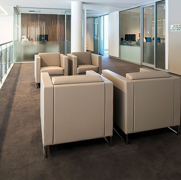 A bright, contemporary commercial lounge or reception area with several cream upholstered armchairs arranged on dark carpet. Frosted and clear glass partitions define adjacent offices or meeting rooms, while a timber feature wall and white structural column add visual contrast.