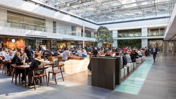 A bright, expansive multi-level commercial atrium functioning as a busy food hall or dining court. Numerous diners sit at timber tables and communal seating around a central illuminated counter, with retail or food-service tenancies lining the perimeter. The space has a glazed roof, extensive upper-level glazing and balconies, tiled flooring, indoor trees and a long translucent floor feature, creating a contemporary urban interior.