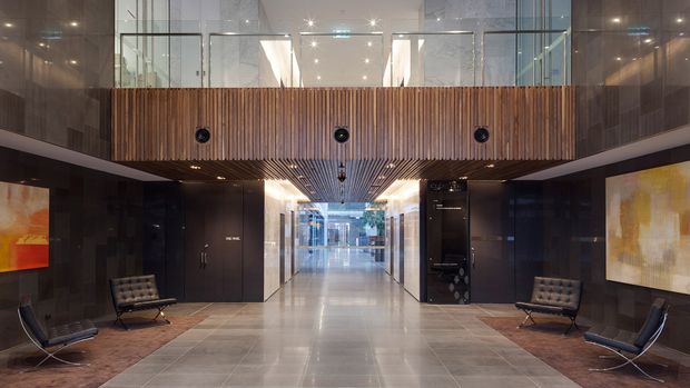A polished double-height commercial lobby or hotel entrance with a central corridor, dark reflective wall panels, large-format grey tiled flooring, and a dramatic timber-slat mezzanine or bridge overhead. Glass balustrades and upper-level glazing create a transparent, contemporary architectural composition. Low lounge chairs sit along both sides, with abstract artwork mounted on the dark walls and recessed lighting throughout.
