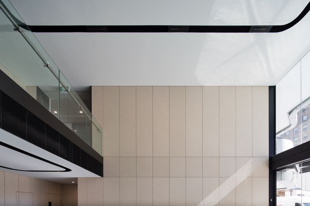 A cropped view of a contemporary double-height interior featuring a tall pale tiled or panelled feature wall, a black-framed glazed mezzanine or upper-level walkway, and a glossy white ceiling with integrated black linear detailing. Strong daylight enters through large black-framed windows on the right, while a sculptural curved black-and-white ceiling element and glass balustrade add a refined modern character.
