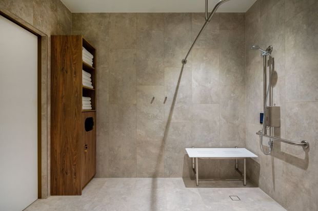 Accessible contemporary bathroom with a large open walk-in shower, handheld and overhead shower fixtures, integrated grab rails, a fold-down shower seat, and full-height beige stone-look wall and floor tiling. A tall dark timber built-in storage unit with open shelving and neatly folded towels is visible beside the doorway.