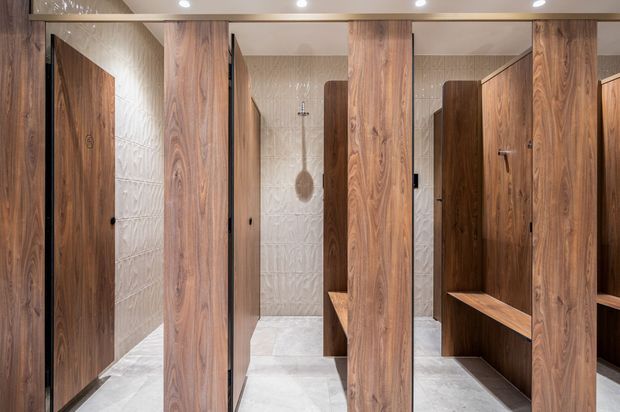A refined commercial-style shower and changing area with multiple timber-look privacy cubicles, integrated benches, pale textured wall tiles, light stone-look flooring, and warm recessed ceiling lighting. The space has a clean, contemporary and upscale appearance.