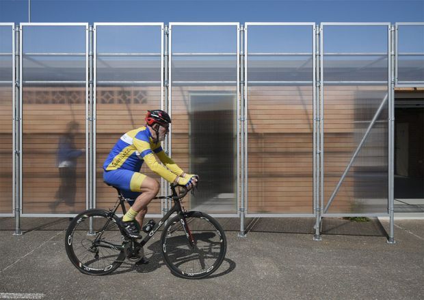 Exterior architectural photograph of a contemporary building facade with a modular translucent and transparent screening system mounted over horizontal timber cladding. A cyclist passes in the foreground, while a dark entry or opening is visible at the right. The facade has a strong repetitive vertical grid and industrial detailing.