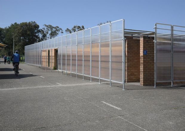 Exterior view of a low-rise institutional or commercial building enclosed by tall translucent screening panels and galvanized metal frames. Exposed brown brick walls and a covered entry or service area are visible, with a paved asphalt forecourt and accessibility signage. The scene is photographed in daylight from outside.
