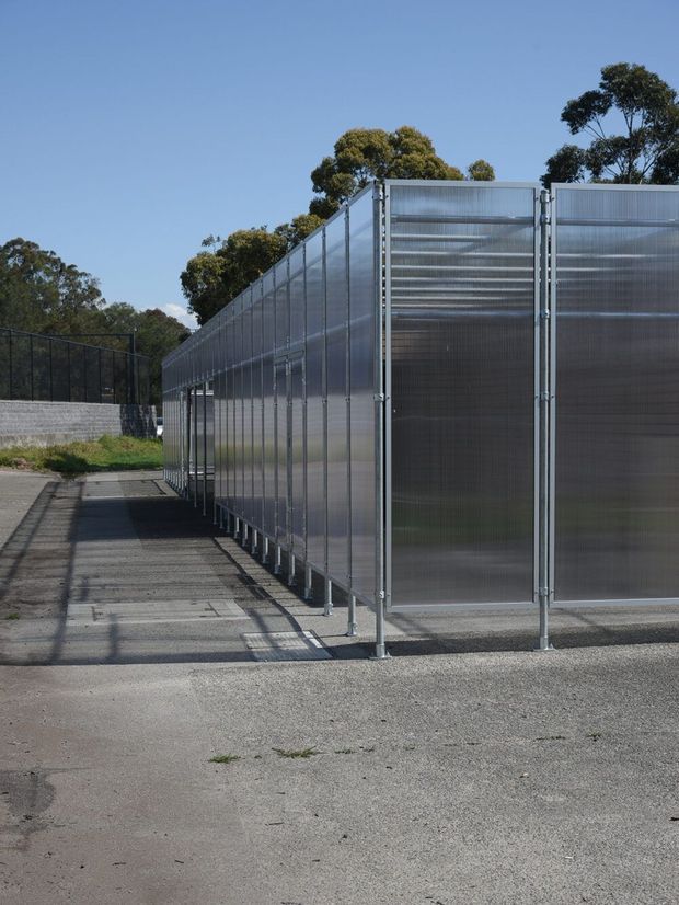 Exterior view of a long, modular enclosure formed by translucent corrugated panels in metal frames, elevated slightly on narrow supports above a paved asphalt surface. The structure appears utilitarian and industrial, with a black mesh fence, low masonry wall, and mature trees in the background under clear daylight.
