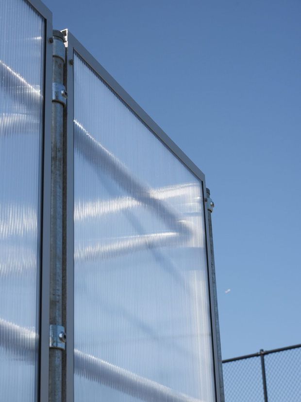 Close-up exterior view of translucent corrugated panel fencing or screening, held in a galvanised metal frame with visible brackets and posts. The panels catch bright sunlight against a clear blue sky, with a chain-link fence in the background. The image focuses on the material, construction, and semi-transparent layered texture rather than an identifiable room or building.