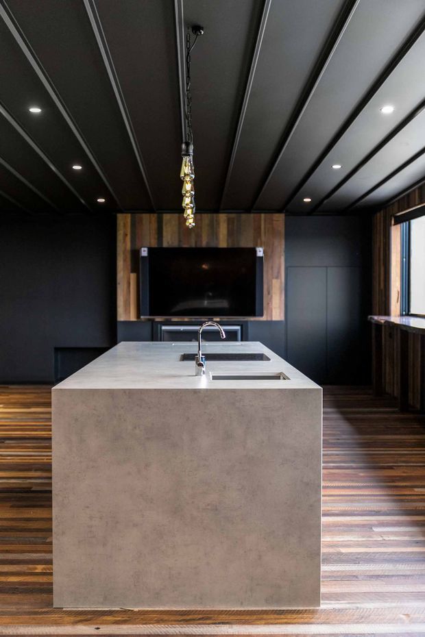 A dark, contemporary kitchen centred on a large monolithic island with an integrated sink and pale concrete-look benchtop. The room features black cabinetry and wall surfaces, a dramatic dark panelled ceiling with recessed downlights, warm reclaimed timber framing around the wall-mounted television, and richly varied timber flooring. A sculptural pendant hangs above the island, while a window and timber-lined wall are visible on the right.