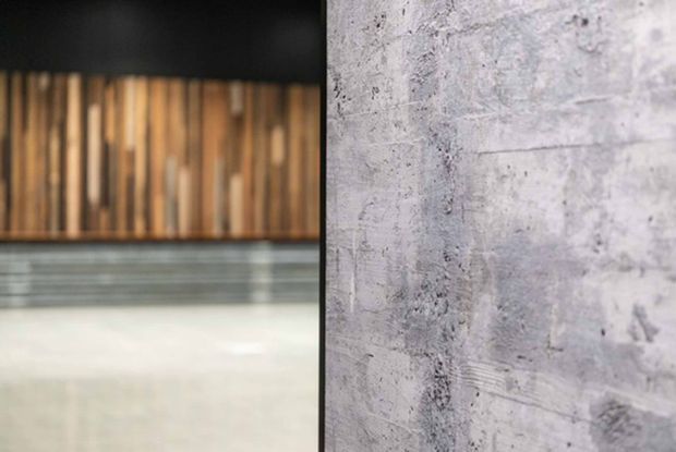 Abstract architectural interior detail showing a rough pale grey concrete wall in the foreground, with a dark ceiling, vertical timber wall cladding, horizontal metal or concrete bands, and a polished light floor receding into the background. The image emphasises contrasting raw concrete and warm timber textures with a minimalist, industrial character.