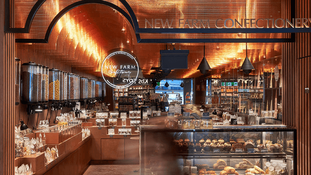 A richly detailed commercial café and confectionery interior with warm copper-toned ribbed wall and ceiling finishes, a long illuminated pastry display counter in the foreground, glass jars and dispensers along the left wall, shelving and retail displays at the rear, and multiple pendant lights over the service area. The space has a sophisticated, industrial-inspired fit-out with strong linear detailing and reflective metal surfaces.