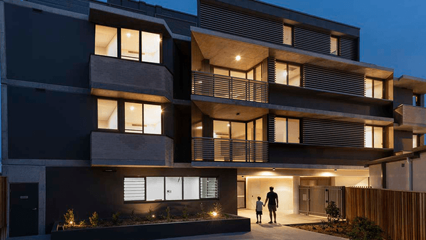Twilight exterior view of a contemporary multi-storey residential building with dark rendered and masonry facades, projecting balconies, horizontal metal balustrades and louvres, warm-lit windows, recessed entry, and a landscaped frontage. Two people are shown approaching the illuminated ground-floor entrance.