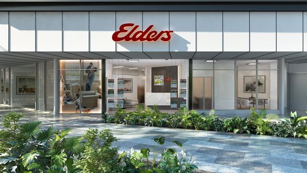 A contemporary commercial building frontage with extensive glass shopfront glazing, light grey panel cladding, and a prominent red Elders sign. The entrance opens onto a landscaped forecourt with tropical planting and a broad paved walkway.
