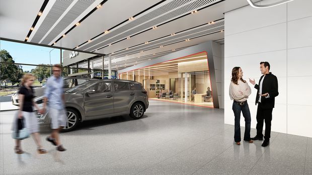 A large contemporary automotive showroom or commercial sales space with a grey vehicle displayed in the foreground, expansive glass frontage, polished light-grey flooring, white wall panels, linear recessed lighting, and a timber-lined retail or reception area beyond. People are present in the showroom, with landscaping visible outside.