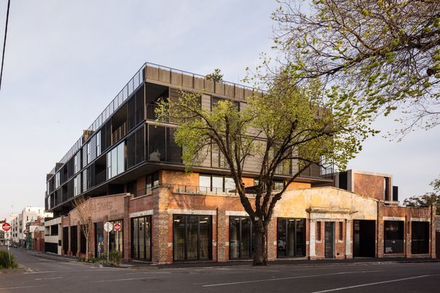 Street-level exterior view of a contemporary multi-storey mixed-use building. The lower level retains and incorporates an aged brick commercial facade with large glazed shopfronts, while the upper levels use dark cladding, extensive glazing, recessed balconies, and rooftop balustrades. The building occupies a prominent urban corner site with mature street trees and surrounding low-rise commercial development.