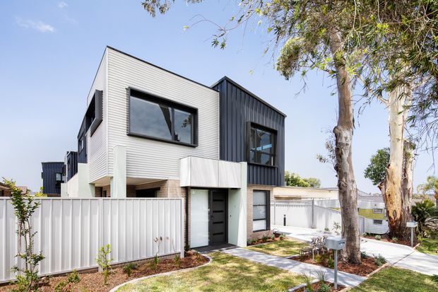 Exterior view of a contemporary two-storey townhouse with contrasting light horizontal cladding and dark vertical cladding, large black-framed windows, a recessed entry, private fencing, and a landscaped front path with mature trees.