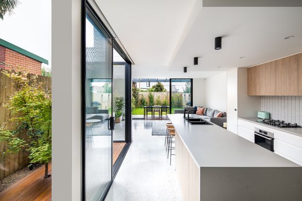 A bright contemporary open-plan kitchen and living area with a large pale island, white cabinetry, warm timber overhead joinery, and a tiled splashback. Full-height black-framed glazing opens toward a landscaped courtyard and dining terrace, while polished light flooring and minimalist black ceiling lights create a clean, airy atmosphere.