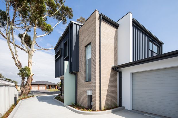 Exterior view of a contemporary multi-storey townhouse or residential dwelling, featuring a light brick central facade, dark vertical metal cladding, white rendered wall planes, narrow vertical glazing, and an attached double garage. A concrete driveway and neighbouring residences are visible, with mature eucalyptus trees along the site.