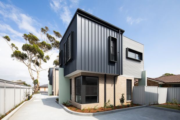 Contemporary multi-level residential townhouse with a strongly articulated projecting upper volume. The facade combines dark vertical metal cladding, pale horizontal cladding, light brickwork, black-framed windows, and pale green rendered side walls. A paved driveway and narrow side access frame the building, with neighbouring residential structures and mature trees visible in the background.