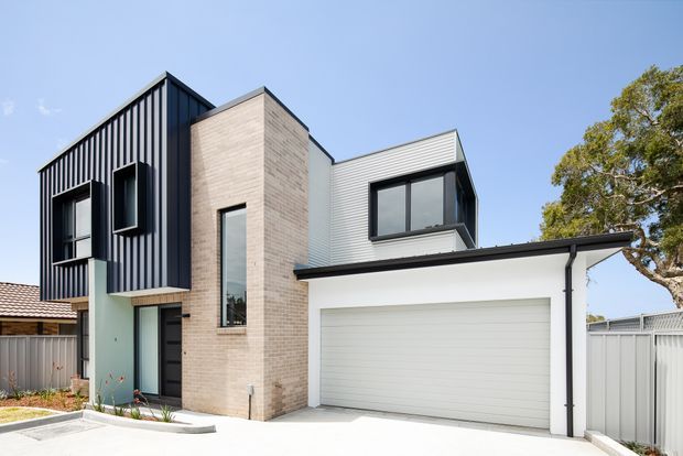 Exterior view of a contemporary multi-level residence with a prominent attached double garage. The facade combines dark vertical cladding, pale brickwork, light corrugated metal upper cladding, expansive dark-framed windows, and a broad low-pitched garage roof. A small landscaped front edge and neighbouring residential structures are visible.