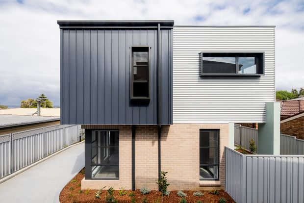 Contemporary two-storey residential exterior with a contrasting dark vertical metal-clad upper volume and light horizontal metal cladding, set above a pale brick ground floor. The facade includes narrow vertical windows, a projecting upper section, and enclosed side fencing around a compact landscaped frontage.