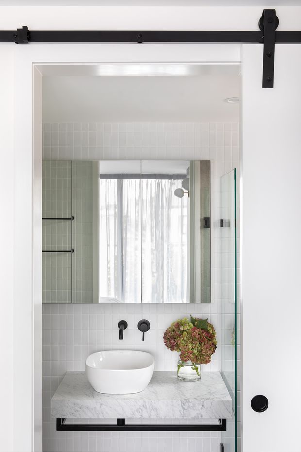 Bright, refined bathroom vanity viewed through a white-framed opening with a black barn-style sliding door track above. The room features white vertical and square wall tiles, a floating marble vanity with a white vessel basin, matte black wall-mounted tapware, a large mirrored cabinet, and a clear glass shower screen. A floral arrangement adds a soft natural accent.
