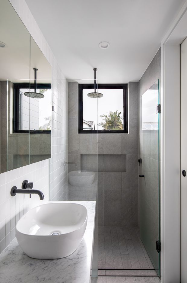 A compact contemporary bathroom with a walk-in glass shower, wall-mounted matte-black fixtures, a white vessel basin on a white marble-look vanity, and large grey wall and floor tiles. A black-framed window brings in natural light, while a full-width mirror reflects the shower area.
