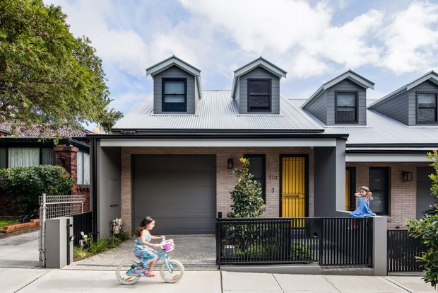 Street-facing view of a contemporary attached townhouse with a light grey metal gabled roof, multiple front dormer windows, pale brick walls, dark grey trims and garage door, and a bright yellow front door. A black metal fence encloses a small front courtyard, with neighbouring attached dwellings visible on either side.