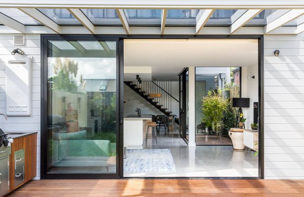 A bright contemporary home exterior viewed from a timber deck, with white horizontal cladding, dark-framed sliding glass doors and a glazed pergola or roof structure. Through the open glazing is a minimalist open-plan interior with a kitchen island, dining area, staircase and courtyard-style greenery. The scene combines indoor-outdoor living with abundant natural light.