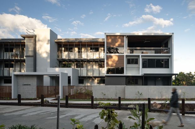 Street-facing view of a contemporary multi-storey residential development with strongly rectilinear massing, recessed balconies, extensive glazing, white rendered walls, dark metal elements, timber-toned screening and a low boundary wall. The image is captured from the landscaped street verge, with pedestrian paths and young planting in the foreground under a bright partly cloudy sky.