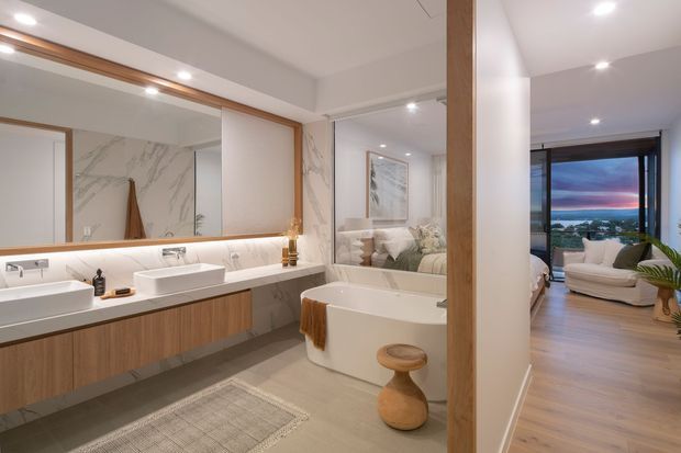 A refined contemporary bathroom with a double vanity, floating timber-look cabinetry, white vessel basins, large marble-look wall panels, and a freestanding white bathtub. Recessed ceiling lighting and a backlit vanity mirror create a bright, polished atmosphere. Through the open passage, a furnished bedroom or sitting area with timber flooring and expansive glazing is visible, along with a distant sunset view.