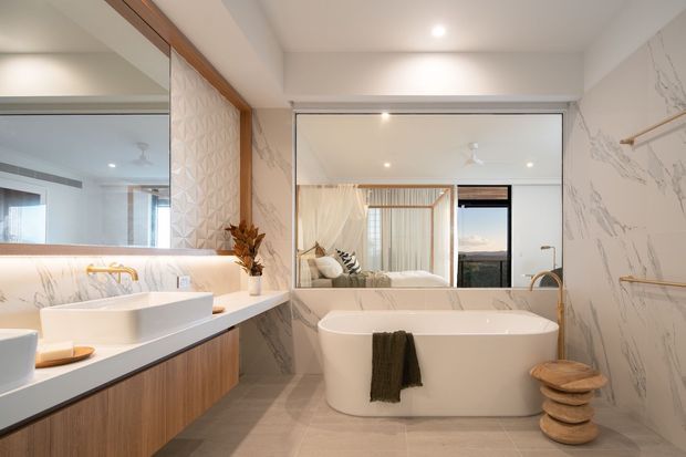 Bright contemporary bathroom with a freestanding white bathtub, double vanity, large mirrored wall, and floor-to-ceiling marble-look wall surfaces. Warm timber vanity cabinetry and trim add contrast, while a bedroom is visible through the mirrored reflection and opening beyond.