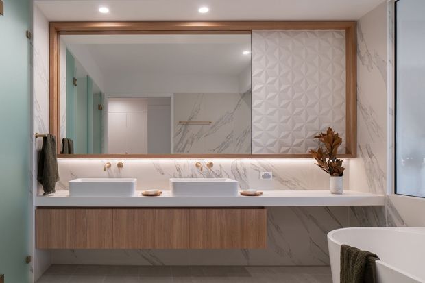 Contemporary double-vanity bathroom with a floating warm-wood vanity, white vessel basins, a pale marble-look wall and benchtop finish, and a large timber-framed mirror. A geometric textured feature panel, recessed downlights, concealed under-mirror lighting, and a partially visible freestanding bathtub create a refined, softly lit fit-out.