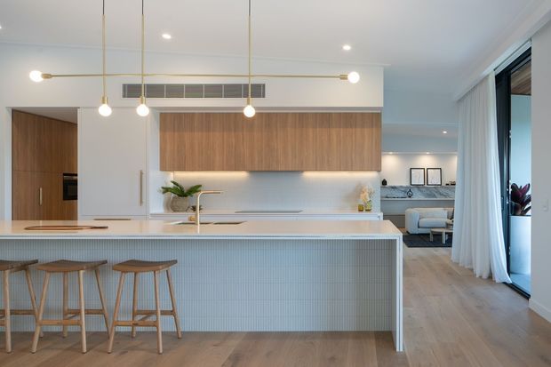 Bright open-plan contemporary kitchen with a large island, pale vertically fluted tiled or panelled front, white benchtops, warm timber cabinetry, and minimalist brass pendant lighting. A timber-floored living area is visible beyond the kitchen, with expansive dark-framed glazing and sheer curtains along the right side.