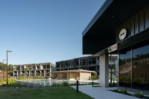Exterior architectural photograph of a contemporary multi-storey residential development arranged around a landscaped courtyard and swimming pool. The complex has strong horizontal and rectilinear forms, extensive glazing, balconies, dark metal roof and soffit elements, pale masonry walls, and a prominent projecting entrance canopy. A sloped, landscaped site and neighboring low-rise buildings are visible in the background.