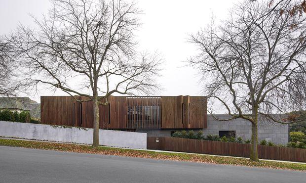 Street-level exterior view of a contemporary residential building set behind a low white rendered wall and dark timber fencing. The facade combines deep brown vertical timber cladding, narrow vertical screening elements, and grey masonry, with mature leafless trees lining the street and partially obscuring the building. The low, elongated form has a flat roof and a restrained modern composition.