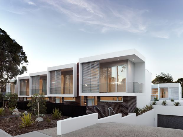 Contemporary multi-level residential development with crisp white cubic forms, extensive floor-to-ceiling glazing, horizontal external blinds, glass balustrades, and dark brick lower-level walls. The homes are set along a landscaped frontage with low white retaining walls and integrated garage access.