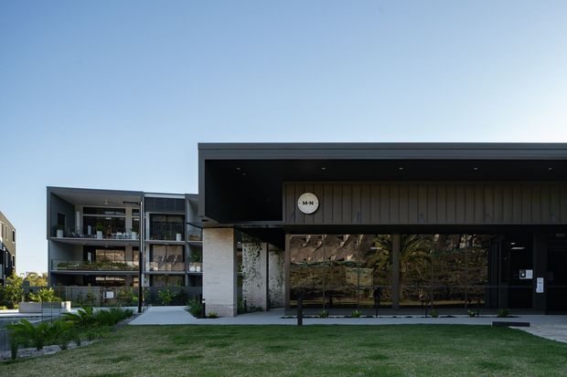 Exterior view of a contemporary multi-storey residential or mixed-use building complex. The foreground building has a broad flat roof canopy, dark metal-looking cladding, large reflective glazed frontage, and a pale masonry or concrete entry column. Apartment balconies and landscaped lawn are visible around the complex.