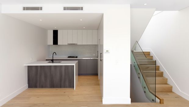 Bright contemporary open-plan kitchen viewed from the living area, with a compact island, integrated pale cabinetry, grey splashback, light timber flooring, and a staircase with timber treads and a clear glass balustrade. The space is minimalist and uncluttered, with white walls and ceiling and abundant natural light.