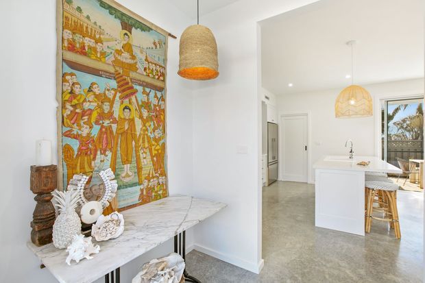 Bright open-plan interior featuring a white kitchen with a central island, bar stools, pendant lighting, pale stone-look flooring, and a marble-topped console. A large colourful figurative wall textile and ornate decorative objects add an eclectic, globally inspired character.
