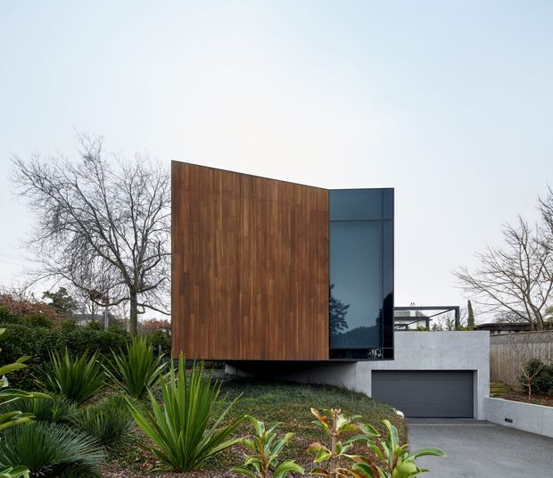 Exterior view of a contemporary residential house set within a landscaped garden. The composition features a large vertical timber-clad upper volume with a tall dark glazed opening, projecting above a rendered concrete or masonry lower level containing an integrated dark garage door. The home has crisp geometric forms, a low-profile roofline, and a restrained palette of timber brown, charcoal, grey, and glass, surrounded by mature deciduous trees and sculptural planting.