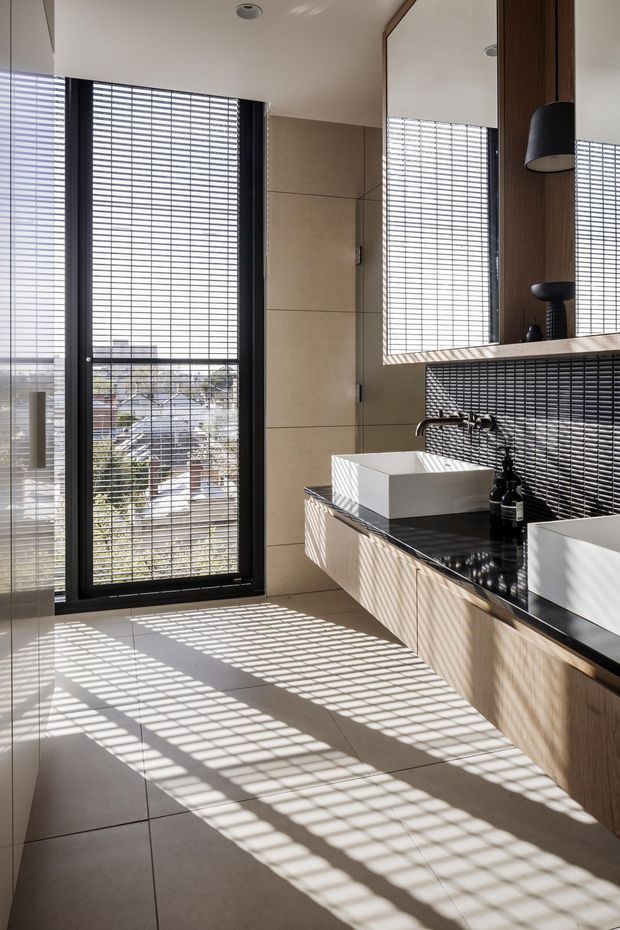 A bright, contemporary bathroom with two white vessel basins set on a dark stone benchtop, integrated timber-look vanity cabinetry, and a dark small-format tiled splashback. A large framed window with horizontal screening casts strong linear shadows across the large neutral floor tiles. A full-height mirror and black wall-mounted pendant lights add depth and contrast.