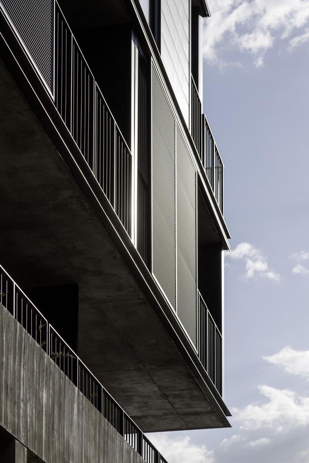 A dramatic upward-looking architectural photograph of a contemporary multi-storey building exterior. Deep cantilevered concrete balconies project from a dark facade, edged with slender black metal balustrades and screened by fine vertical or perforated metal panels. Strong geometric lines and deep shadows contrast with the bright partly cloudy sky.