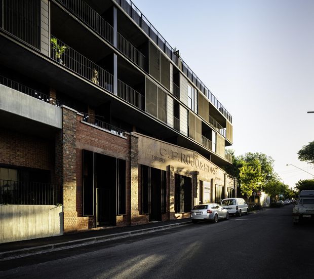Street-level exterior view of a contemporary multi-storey apartment or mixed-use building. The lower facade combines exposed red brick and dark recessed openings, while the upper levels feature projecting balconies with slender dark metal railings, concrete elements, and vertical screening. The building fronts an urban residential street with parked cars and mature trees.